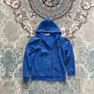 Blå hoodie från Comme des Garçons - Snygg blå hoodie med dragkedja från Comme des Garçons. Har en huva, två fickor fram och det ikoniska svarta hjärtat med ögon på bröstet och ryggen. Perfekt för dig som gillar streetwear och vill sticka ut med en unik detalj.