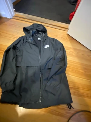 Svart Nike vindjacka med huva - Svart vindjacka från Nike med dragkedja och huva. Jackan har en vit Nike-logga på bröstet och är tillverkad i lätt, vattenavvisande material. Perfekt för blåsiga dagar och enkel att packa ner. Justerbar nederkant med dragsko för bättre passform.