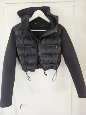 Svart cropped pufferjacka med huva,stl.M, Zara - Snygg svart cropped pufferjacka från Zara, med stor huva och dragsko i midjan. Jackan har quiltad front och rygg i glansigt material samt släta ärmar i mjuk tyg. Perfekt för våren och hösten.Jackan är bara 36-38 cm lång,  och ger en cool streetstyle-look.
