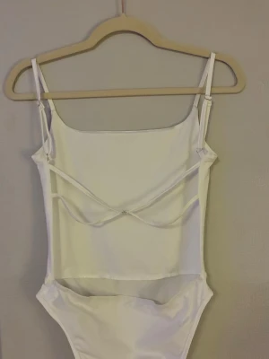 Vit body med öppen rygg  - Snygg vit body med tunna, justerbara axelband och raka linjer. Bodyn har en unik detalj baktill med extra band över ryggen och är tillverkad i ett stretchigt, slätt material som ger en tight passform. Perfekt att styla med jeans eller kjol.