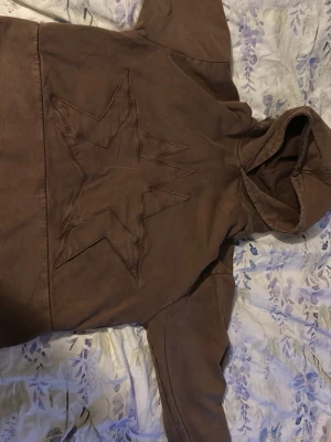 Brun hoodie med stjärndetalj - Säljer en brun hoodie med stor stjärndetalj sydd på framsidan och en liten stjärna broderad i nacken. Hoodien har en rymlig huva och är gjord i ett mjukt bomullsliknande material. Perfekt för dig som gillar streetwear och unika detaljer.