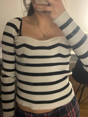 Randig ribbad topp med sweetheart-ringning - Säljer en vit ribbad topp med svarta horisontella ränder och lång ärm. Toppen har en snygg sweetheart-ringning och figurnära passform. Perfekt för dig som gillar trendiga och bekväma plagg med lite edge.