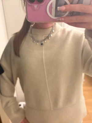 Beige stickad tröja med ribbade detaljer - En enkel och stilren beige stickad tröja med rund hals och ribbade muddar vid ärmslut och nederkant. Tröjan har en diskret söm framtill som ger en modern touch. Perfekt att matcha med jeans eller shorts.