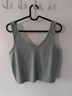 Grågrön ribbad linnetopp - Snygg grågrön ribbad linnetopp med breda axelband och v-ringad hals både fram och bak. Toppen är croppad och har en enkel, stilren look som passar perfekt till jeans eller kjol. Materialet känns mjukt och stretchigt för en bekväm passform.