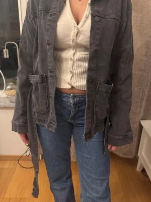 Svart jeansjacka med knytskärp - Cool svart jeansjacka med knytskärp i midjan och stora fickor framtill. Jackan har klassisk krage, knäppning med knappar och avslappnad passform. Perfekt för dig som gillar en lite oversized look och vill ha en unik twist på den klassiska jeansjackan.