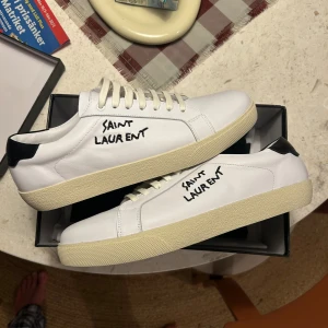 Saint Laurent skor  - Snygga vita sneakers från Saint Laurent med svart detalj på hälen och handskriven logga på sidan. Skorna har klassisk låg profil, vita snören och en beige sula. Tillverkade i läder för en lyxig känsla och stilren look.