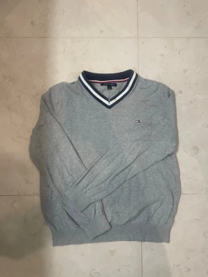 Grå v-ringad tröja Tommy Hilfiger - Snygg grå v-ringad tröja från Tommy Hilfiger med klassisk logga broderad på bröstet. Tröjan har ribbade muddar och en krage med vita och marinblå detaljer. Perfekt för en stilren och avslappnad look. Passar mer S 