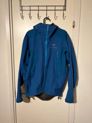 Arc'teryx regnjacka  - Säljer en blå regnjacka från Arc'teryx med huva och hel dragkedja framtill. Jackan har två sidofickor med dragkedja och logotypen är broderad på bröstet. Materialet är lätt och vattenavvisande, perfekt för regniga dagar. Jackan har justerbar nederkant och ärmar.