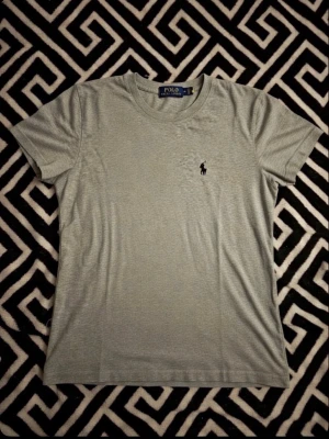 Grå t-shirt från Polo Ralph Lauren - Snygg grå t-shirt från Polo Ralph Lauren med klassisk rund hals och ikoniska broderade loggan på bröstet. Tillverkad i mjuk bomull för skön passform. Perfekt basplagg till garderoben.