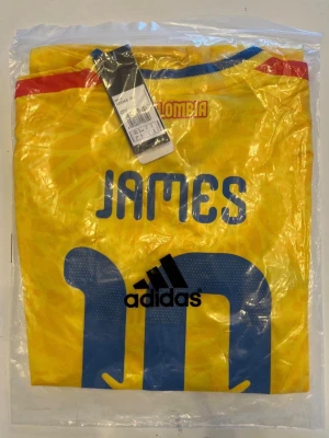 Colombia fotbollströja 2026 VM  - Helt ny! Gul Colombia fotbollströja från Adidas med blå detaljer och tryck 'JAMES' samt nummer 10 på ryggen. Tröjan har blå och röda inslag vid halsen och är tillverkad i ett lätt, ventilerande material perfekt för sport.