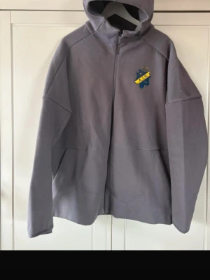 Grå AIK hoodie från Adidas - (Pris går att diskuteras) Snygg grå hoodie från Adidas med AIK-emblem på bröstet och Adidas-logga på ärmen. Tröjan har dragkedja framtill, huva och är tillverkad i ett mjukt, sportigt material. Perfekt för dig som vill representera AIK med stil.