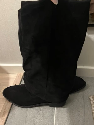 Svarta höga mockastövlar - Säljer ett par svarta höga stövlar i mjuk mocka. De har rundad tå, låg klack och en slouchy passform som ger en avslappnad men ändå stilren look. Perfekta till både jeans och kjol för en trendig outfit.