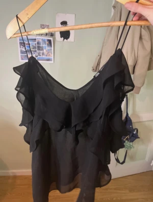 Svart volang topp från Zara - Säljer en svart, tunn och luftig topp från Zara med smala axelband och flera lager volanger framtill.Materialet känns lätt och svalt, toppen är ärmlös och har en snygg v-ringning.