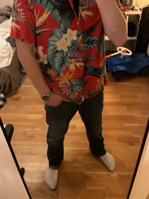 Röd blommig hawaiiskjorta  - Storlek L men passar snarare s eller m, skriv för mer information.Färgstark hawaiiskjorta från pull&bear med röd bas och stora blommiga mönster i blått, vitt, orange och grönt. Skjortan har korta ärmar, klassisk krage och knappar framtill. Perfekt för dig som vill sticka ut med en tropisk vibe.