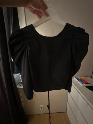 Svart top med puffärmar - Trendig svart top med stora puffärmar och öppen rygg med snygga knytband. Toppen har rund halsringning och är tillverkad i ett slätt tyg som ger en stilren look. Perfekt för dig som vill sticka ut med en cool och modern stil.