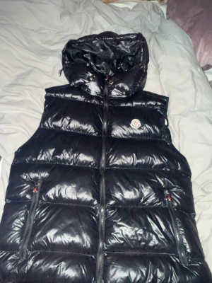 Svart dunväst från Moncler med luva - Snygg svart dunväst från Moncler med glansig finish och avtagbar luva. Västen har två dragkedjefickor fram och klassisk Moncler-logga på bröstet. Perfekt för lager på lager och riktigt trendig look.