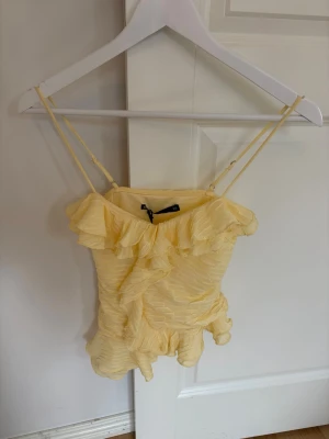 Gul volangtop från Bik Bok - Supersöt ljusgul topp från Bik Bok med volangdetaljer och smala axelband. Toppen har en feminin och lekfull look med rynkade detaljer och är perfekt för varma dagar. Materialet är mjukt och lätt, vilket gör den extra bekväm.