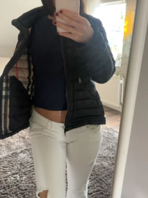 Svart pufferjacka från Burberry - Säljer en svart pufferjacka från Burberry med klassiskt rutigt foder på insidan. Jackan har dragkedja framtill och två sidofickor med dragkedja. Perfekt för kalla vinterdagar och riktigt snygg till alla outfits.