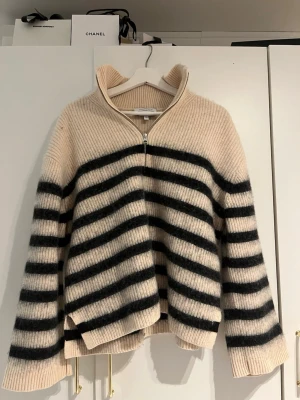 Beige stickad tröja med svarta ränder - Säljer en beige stickad tröja med svarta horisontella ränder och halvzip framtill. Tröjan har hög krage och är ribbstickad med breda ärmar. Materialet ser ut att vara mjukt och fluffigt, perfekt för att hålla sig varm. Märket är H&M.