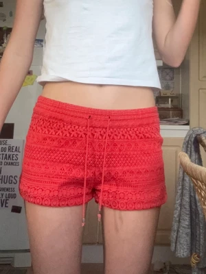 Vera Moda-coral spets shorts - Snygga coral shorts med virkad spets och snörning i midjan. De har en feminin volangkant och är perfekta för varma sommardagar. Shortsen har en relaxed fit och elastisk midja för extra komfort.