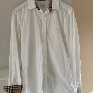 Vit Burberry skjorta  - Klassisk vit skjorta från Burberry med ikoniska rutiga detaljer på insidan av kragen och ärmsluten. Skjortan har långa ärmar, knappar framtill och diskret broderad logga på bröstet. Perfekt för en stilren och tidlös look. Nypris: 4000 kr, mitt pris: 850