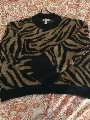 Zebramönstrad stickad tröja H&M - Cool stickad tröja från H&M med zebramönster i svart och brun. Tröjan har rund halsringning, långa ärmar och ribbade muddar vid ärmslut och nederkant. Perfekt för dig som vill sticka ut med ett djurmönster i garderoben.