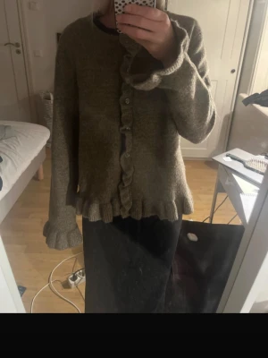 Brun volangkofta med pärlknappar - Mysig brun kofta med volangdetaljer längs knäppningen, ärmsluten och nederkanten. Koftan har pärlknappar framtill och är stickad i ett mjukt material. Perfekt för lager på lager och ger en söt vintagekänsla till din outfit.