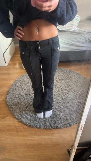 Mörkgrå bootcut jeans med fickor - Säljer ett par mörkgrå bootcut jeans med låg midja och dekorativa fickor fram. Jeansen har breda hällor och vita knappar som detaljer. Perfekta för dig som gillar en retro vibe och vill ha ett par snygga jeans till vardagslooken.