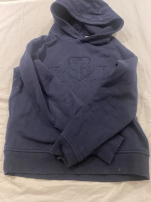 Mörkblå hoodie från GANT - Mörkblå hoodie från GANT med broderad logga och texten 'American Sportswear' framtill. Klassisk huva och långa ärmar, perfekt för en avslappnad och sportig stil. Mjuk och skön känsla tack vare bomullsmaterialet.
