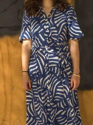 H&M Blåvit mönstrad skjortklänning - Säljer en H&M blå skjortklänning med vitt abstrakt mönster och knappar framtill. Klänningen har korta ärmar, krage och ett matchande knytband i midjan för en snygg siluett. Perfekt för dig som gillar statement-plagg med coola prints. Storlek XL. Perfekt skick