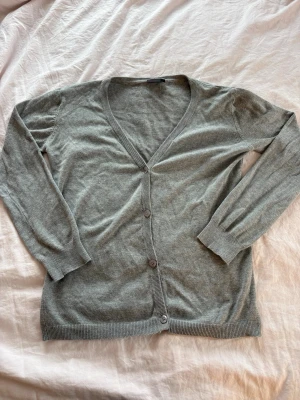 Ljusgrå basic kofta med knappar - En enkel och stilren ljusgrå kofta med v-ringning och tre knappar framtill. Koftan har långa ärmar och ribbade muddar vid ärmslut och nederkant. Perfekt att slänga över en t-shirt för en chill look.