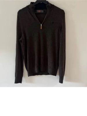 Mörkbrun halvzip-tröja från Morris  - Mörkbrun stickad tröja från Morris med halv dragkedja och liten broderad logga på bröstet. Tröjan har ribbade muddar vid ärmslut och nederkant, samt en klassisk passform. Perfekt att bära över en t-shirt eller skjorta.
