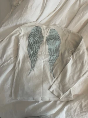 Vit långärmad topp med änglavingar - Vit långärmad topp från Ember med tryck av detaljerade änglavingar i grått på ryggen. Klassisk rund halsringning och mjukt bomullsmaterial. Perfekt för dig som gillar unika detaljer på dina basplagg.