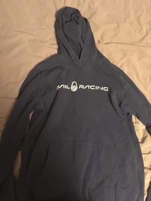 Blå hoodie från Sail Racing - Mörkblå hoodie från Sail Racing med vit logga tryckt över bröstet. Klassisk känguruficka framtill och stor huva. Tillverkad i mjukt bomullsmaterial som är skönt mot huden. Perfekt för en avslappnad och sportig stil.