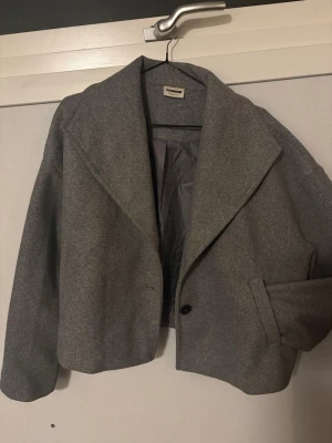 Grå kappa från Vero Moda  - Stilren grå kappa från Vero Moda med bred krage och en knapp framtill. Kappan har en rak, boxig passform och är fodrad på insidan. Perfekt för dig som gillar minimalistisk och modern stil. Materialet känns som ullmix.