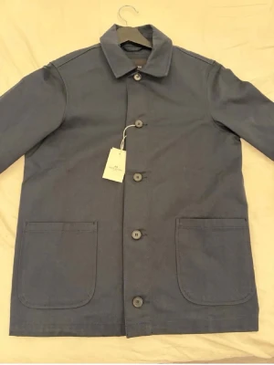 Mörkblå overshirt från John Henric - Stilren mörkblå overshirt från John Henric som har helt ny med prislapp kvar! Med klassisk krage, stora knappar och två rejäla framfickor. Tillverkad i ett kraftigt bomullstyg som ger en clean och modern look. Perfekt att slänga över en hoodie eller t-shirt. 