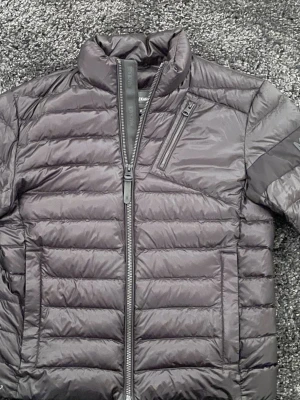 Svart dunjacka från Mackage  - Snygg svart dunjacka från Mackage  med quiltad design, hög krage och dragkedja framtill. Jackan har en bröstficka med dragkedja och diskret logga på ärmen. Perfekt för kalla vinterdagar och riktigt stilren look.