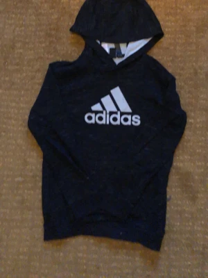Svart hoodie från Adidas med logga - Snygg svart hoodie från Adidas med stor vit logga på bröstet. Klassisk modell med huva och ribbade muddar. Perfekt för en sportig och avslappnad stil. Materialet känns mjukt och bekvämt mot huden.