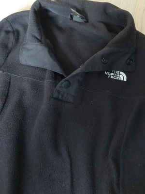 Svart fleece från The North Face - Säljer en svart fleece med hög krage och tryckknappar från The North Face. Tröjan har broderad logga på bröstet och är gjord i mjukt fleecematerial. Perfekt för lager-på-lager och håller dig varm under kyliga dagar.