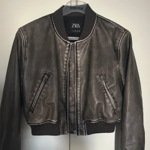 Bomberjacka från ZARA - Snygg grå/svart bomberjacka från ZARA med glansig finish och ribbade muddar. Jackan har två snedställda fickor med dragkedja och klassisk bomberkrage. Perfekt för dig som vill ha en stilren och trendig look. Inga defekter.