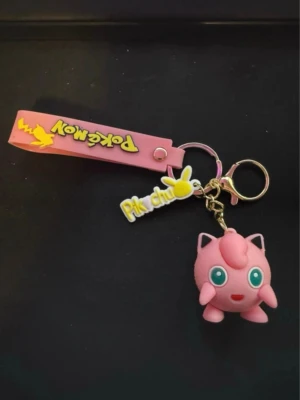 Pokémon nyckelring med Jigglypuff - Säljer en söt nyckelring med en rosa Jigglypuff-figur och Pikachu-detaljer. Nyckelringen har även ett rosa band med Pokémon-logga och Pikachu-tryck. Perfekt accessoar för Pokémon-fans som vill piffa till sina nycklar eller väskor.