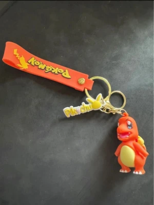 Pokémon nyckelring med Charmander - Cool nyckelring med en orange Charmander-figur i mjukt gummi, gul Pikachu-tag och röd Pokémon-rem. Perfekt accessoar för Pokémon-fans som vill pimpa sina nycklar eller väskor.