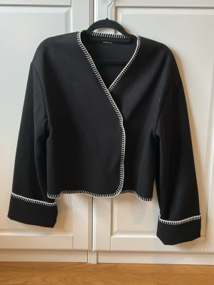 Svart cardigan med vit kontrastsöm - Stilren svart cardiganjacka från Gina Tricot med vit kontrastsöm längs kanterna och ärmsluten. 