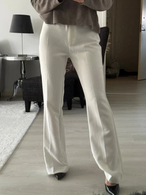 Vita kostymbyxor med pressveck - Säljer ett par stilrena vita kostymbyxor med raka ben och pressveck framtill. Byxorna har hög midja och är tillverkade i ett mjukt, vävt material som ger en snygg siluett. Perfekta för en clean och modern look.