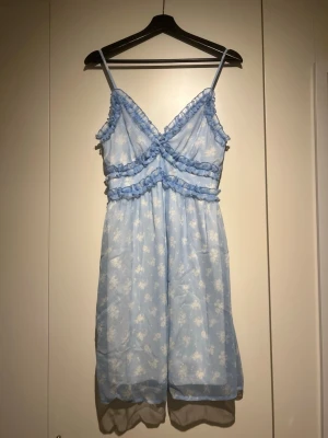 Ljusblå blommig klänning med volang - Söt ljusblå klänning med vita blommor och volangdetaljer. Klänningen har smala axelband, omlott framtill och är kort i modellen. Perfekt för sommarens varma dagar och har en luftig känsla.