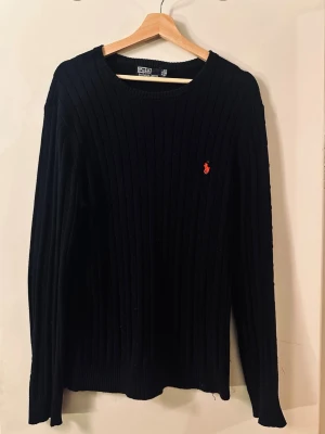 Svart stickad tröja från Polo Ralph Lauren - Klassisk svart stickad tröja från Polo Ralph Lauren med rund halsringning och ribbade detaljer. Tröjan har det ikoniska röda Polo-märket broderat på bröstet och är tillverkad i ett mjukt, stretchigt material. Perfekt till jeans eller kjol.