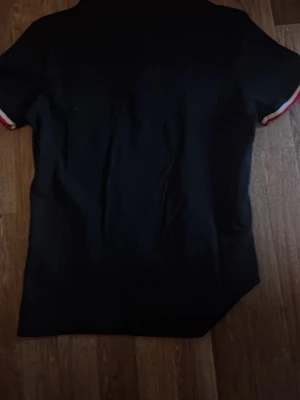 Svart pikétröja med rödvit krage - Snygg svart pikétröja med krage och knappar framtill. Kragen och ärmsluten har röda och vita detaljer som ger en sportig look. Liten logga på bröstet. Perfekt till jeans eller shorts.