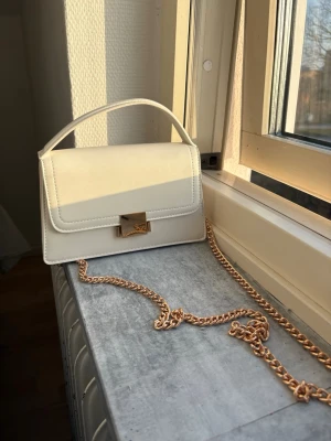 Vit väska med guldkedja - Snygg vit handväska i boxig modell med guldfärgad kedja och metallspänne framtill. Väskan har ett stilrent och modernt utseende med ett kort handtag upptill. Perfekt accessoar för att lyfta din outfit. Se sista bild för defekt bara en liten fläck inuti. 