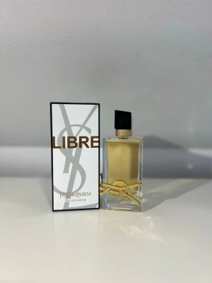 Yves Saint Laurent Libre Eau de Parfum - YSL Libre Eau de Parfum med elegant rektangulär glasflaska, guldiga detaljer och svart asymmetrisk kork. Doften kommer i en stilren vit och guldig förpackning med stor YSL-logga. Perfekt för dig som gillar lyxiga och moderna parfymer.