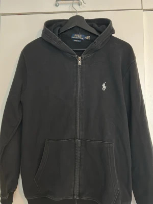 Svart hoodie från Polo Ralph Lauren - Säljer en svart hoodie från Polo Ralph Lauren med dragkedja framtill och klassisk huva. Tröjan har två stora fickor fram och den ikoniska vita loggan broderad på bröstet. Tillverkad i mjuk bomull för skön passform och stilren look. Desvärre saknas snören men är lätt o billigt att köpa nya
